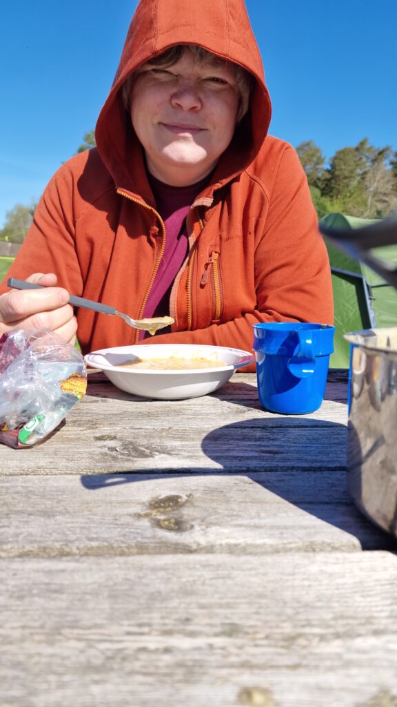 jag i en orange luvjacka med en knallblå himmel äter ärtsoppa vid ett träbord